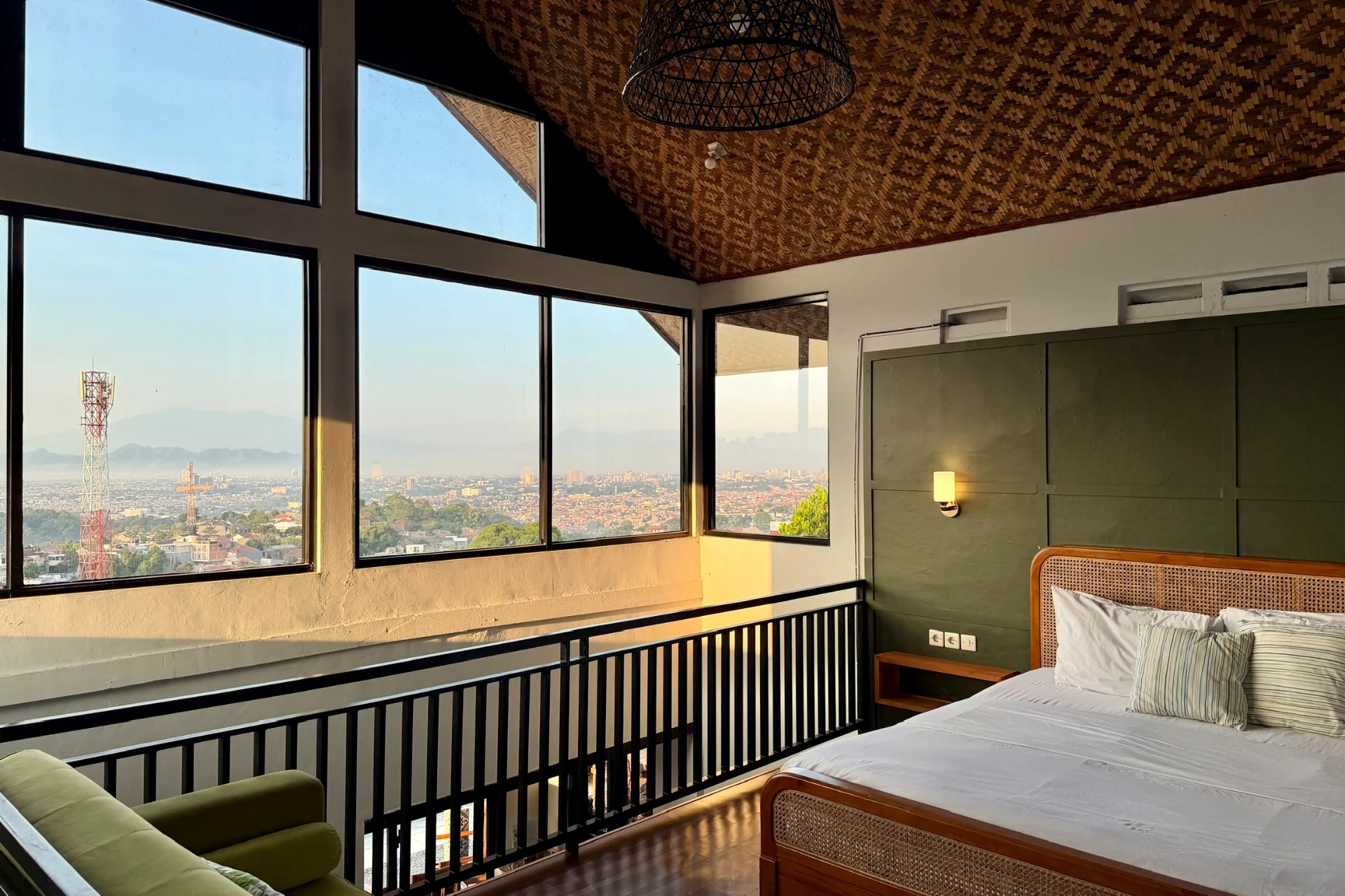 Queen bed Kamar 1 di samping jendela dari lantai ke plafon dengan langit-langit rotan anyaman dan pemandangan Bandung saat matahari terbit