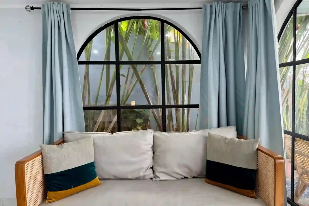 Jendela lengkung dengan gorden biru muda dan sofa rotan di sudut baca Kamar 4