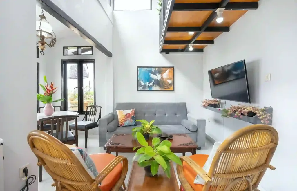 Ruang keluarga Villa Yucca dengan sofa bed abu-abu, kursi rotan bantal oranye, meja kopi kayu ukir, dan smart TV