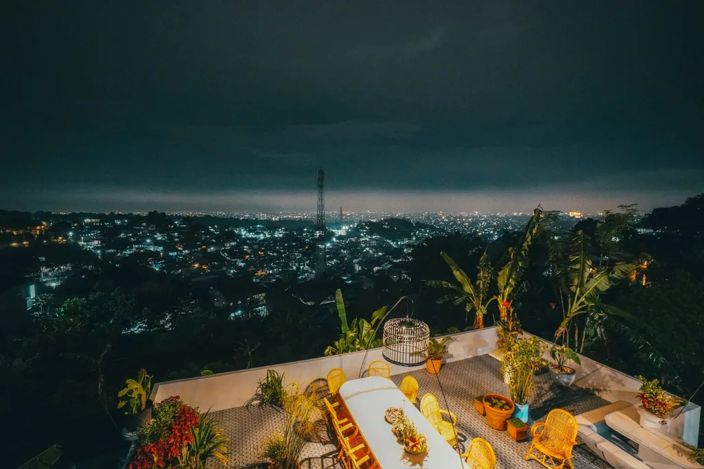 Pemandangan malam hari lampu kota Bandung dari teras atas Villa Yucca dengan kursi rotan kuning