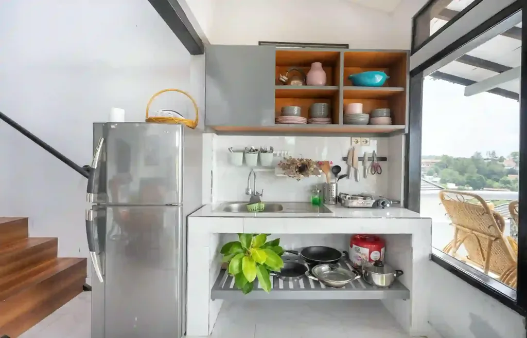 Kitchenette ringkas dengan meja marmer, kompor gas, kulkas stainless, dan rak terbuka berisi peralatan makan di samping balkon